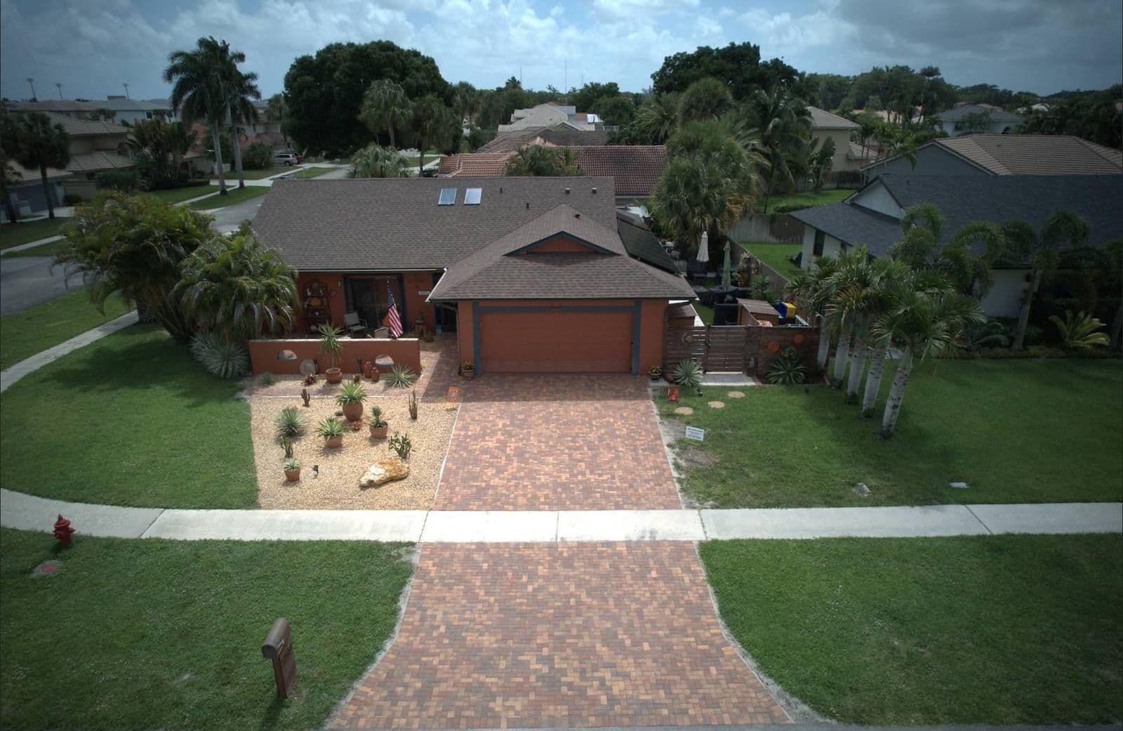 Aerial view of a single-story red brick house with a two-car garage, brick driveway, and landscaped yard with palm trees in a residential Florida neighborhood