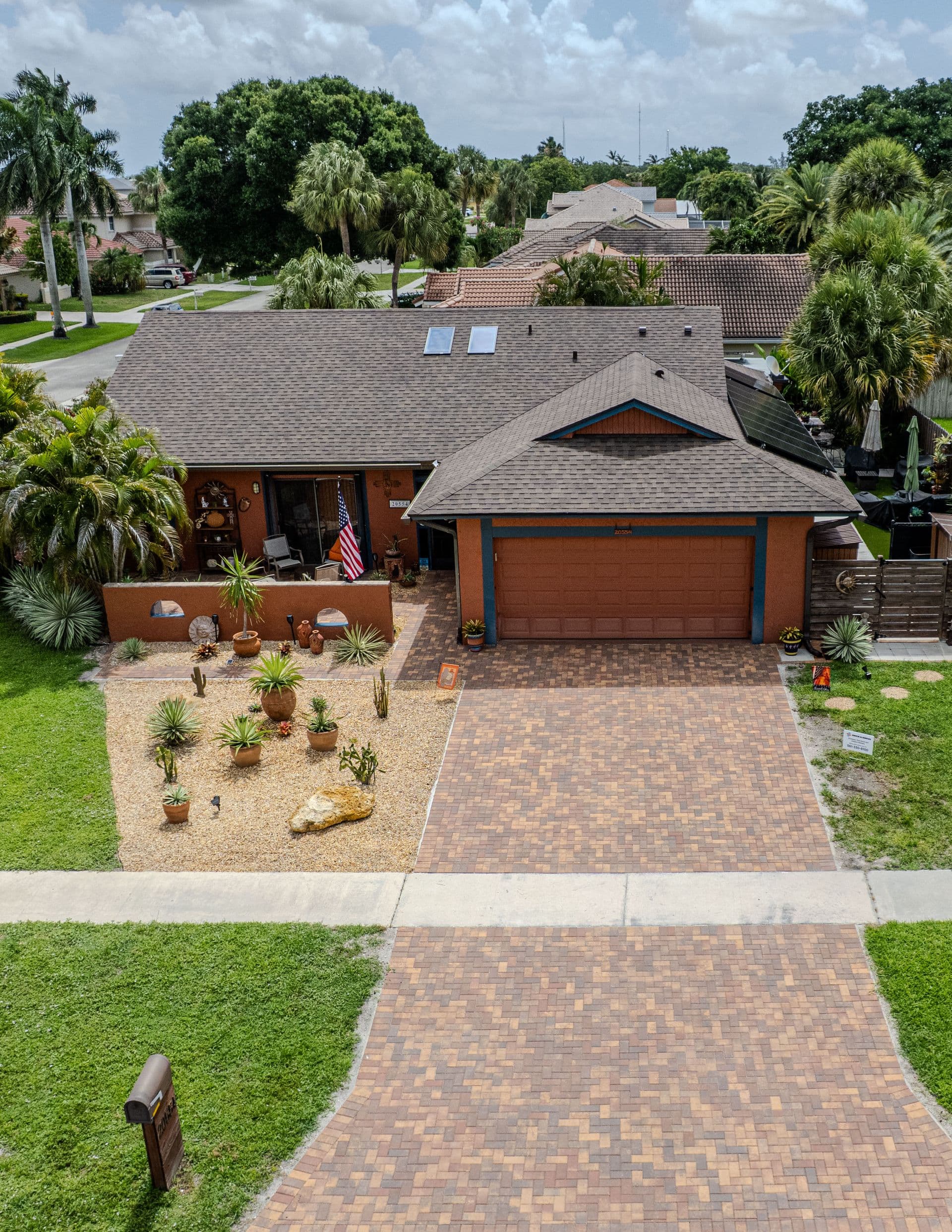 Orange and blue single-story residential home with brick driveway, desert landscaping with cacti and palm trees, and two-car garage in suburban neighborhood