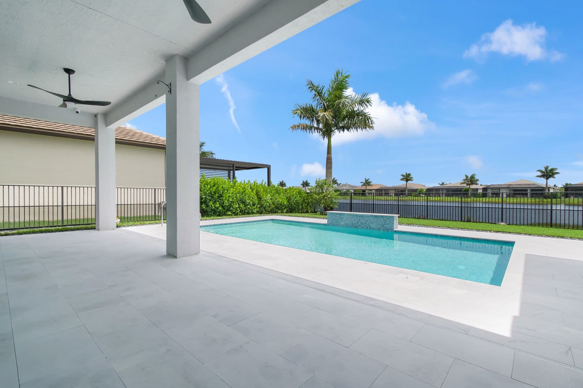Modern backyard oasis with covered patio, rectangular swimming pool, palm trees, and manicured lawn overlooking water feature and residential community