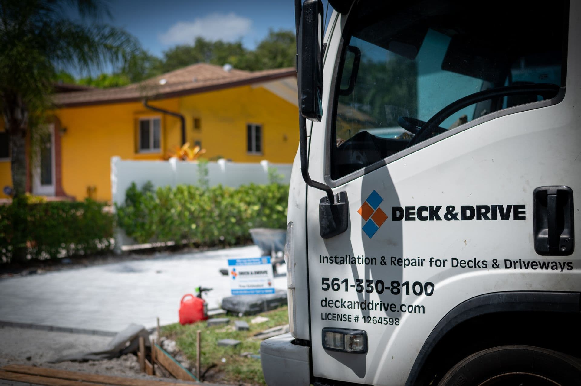 White Deck & Drive truck advertises deck and driveway installation and repair beside a sunny yellow home, hedge, and concrete driveway.