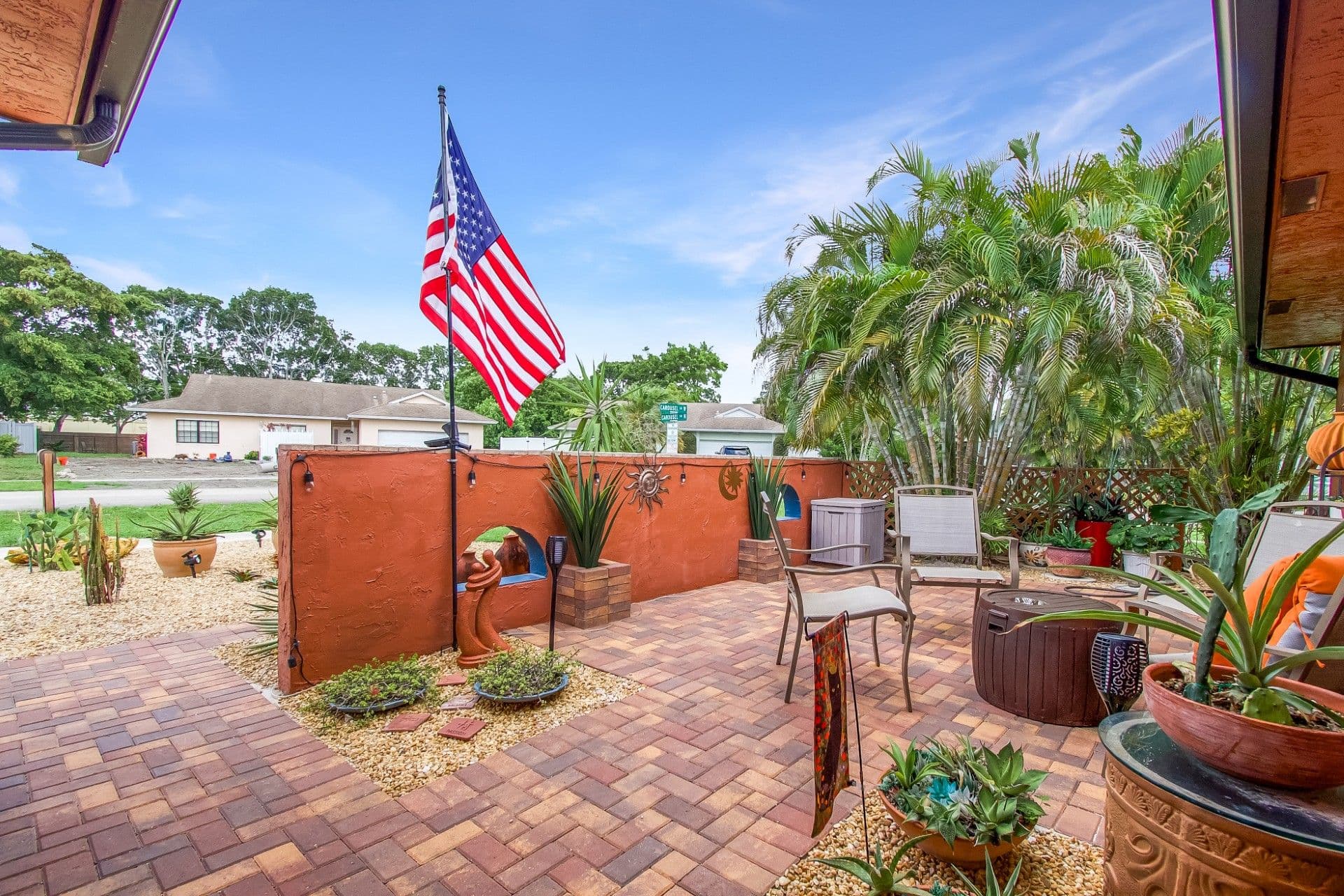 Backyard patio with red brick pavers, rust-colored decorative wall structure, American flag, potted tropical plants including palm trees, and outdoor seating furniture