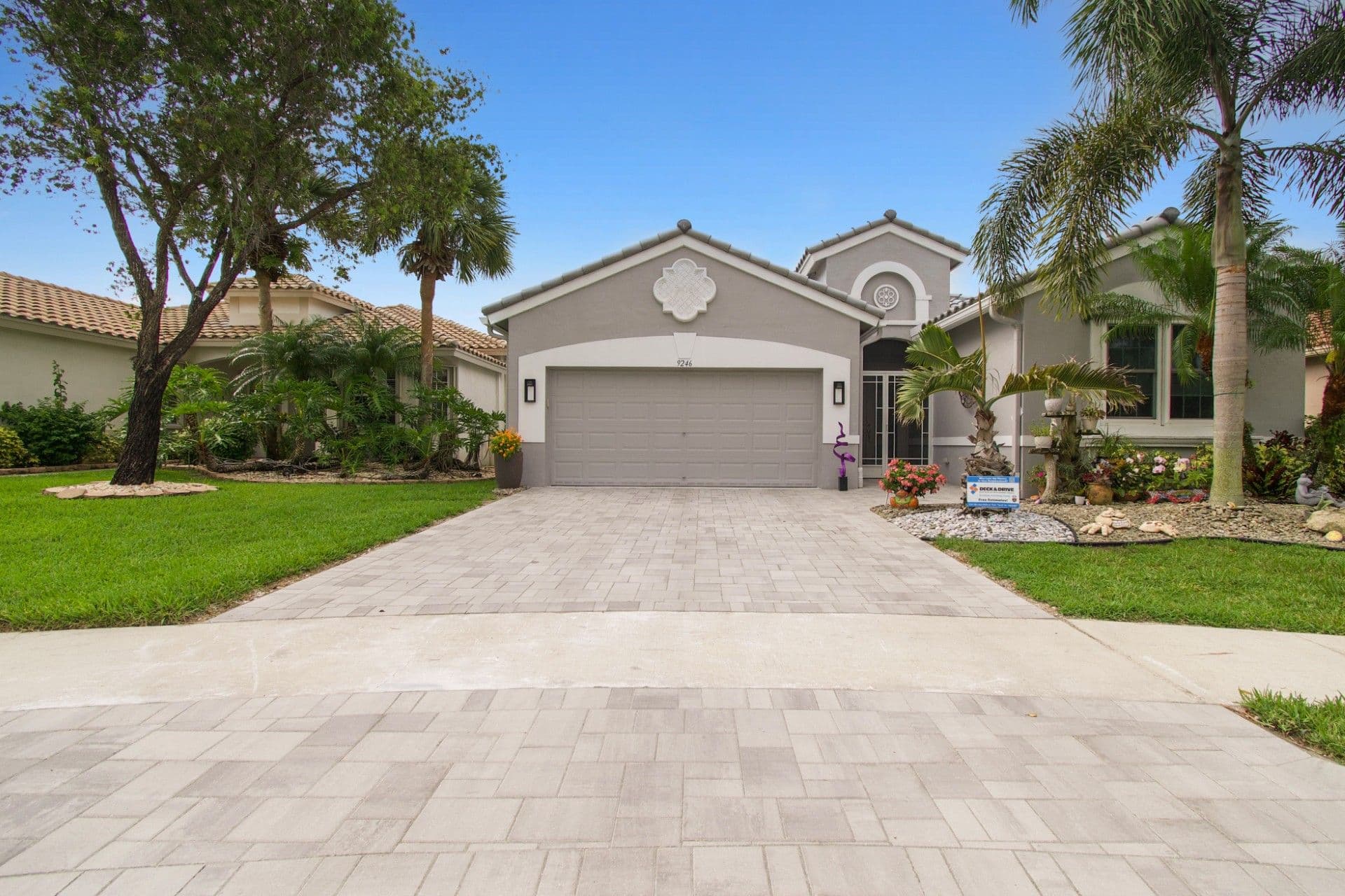 Single-story beige stucco house with gray roof, two-car garage, paver driveway, and tropical landscaping including palm trees and flowering plants